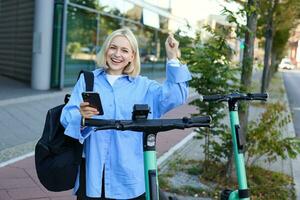 Porträt von jung lächelnd weiblich Modell, mieten ein elektrisch Roller, mit Handy, Mobiltelefon Telefon zu Scan qr Code mit Smartphone Anwendung, Reiten Zuhause von Universität, Stehen draußen auf Straße foto