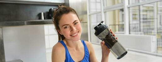 lächelnd Fitness Mädchen Getränke Wasser im das Mitte von Ausbildung Sitzung, lächelnd und suchen Glücklich, trainieren von Zuhause im Blau sportbra foto