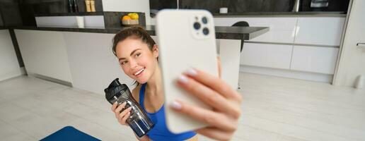 Porträt von Sportlerin nimmt Selfie mit Wasser Flasche im Leben Zimmer, hält Smartphone und posiert zum Foto während tun Fitness trainieren