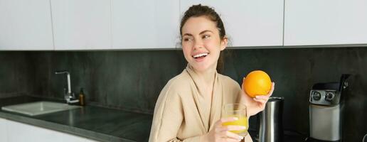 Porträt von gut aussehend Brünette Frau, Trinken frisch Orange Saft im Küche, halten Obst im Hand, Lachen und lächelnd, suchen glücklich foto