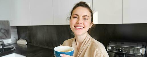 Lebensstil Konzept. Porträt von glücklich Brünette Frau im Bademantel, Trinken Kaffee im das Küche, haben Morgen Tasse Tee und lächelnd foto