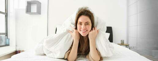 süß Mädchen mit unordentlich Haar, Lügen im Bett bedeckt im Weiß Blätter Bettdecke, lächelnd und Lachen kokett, Ausgaben Zeit im ihr Schlafzimmer foto