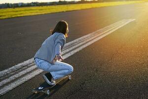 Seite Aussicht von schön asiatisch Mädchen auf Skateboard, Reiten ihr Kreuzer gegenüber das Sonne auf ein leeren Straße. glücklich jung Skater genießen sonnig Tag auf ihr Schlittschuh foto