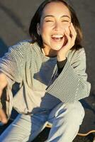 Selfie von schön asiatisch Mädchen lächelnd, nehmen Foto auf Smartphone während Sitzung auf Skateboard draußen