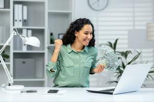 online Kasino. ein jung Muslim Frau sitzt im das Büro beim ein Tabelle mit ein Laptop, hält Kasse Geld im ihr Hand, Theaterstücke online Spiele, setzt Wetten, feiert Gewinn. foto