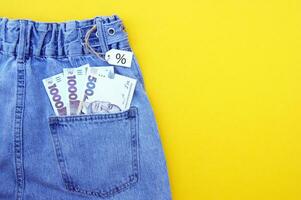 eben legen, Detail von Denim Jeans mit ukrainisch Griwna Rechnungen im Tasche mit Weiß Etikette auf Gelb Hintergrund. foto