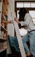 Vertikale Aufnahme einer südasiatischen jungen Frau auf der Treppe, die ein Buch aus einem Bücherregal auswählt foto