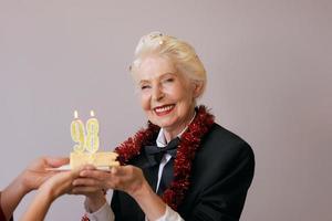 Fröhliche, stilvolle, achtundneunzig Jahre alte Frau im schwarzen Anzug, die ihren Geburtstag mit Kuchen feiert. Lifestyle, Positiv, Mode, Stilkonzept