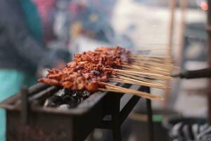 Hähnchen Satay Über traditionell Holzkohle Feuer. das Satay brennt mit Rauch und ein appetitlich Aussehen. Hände Kochen Satay auf das Grill. Menschen sind schon Schlange stehen. foto