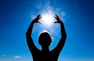 Silhouette von ein Mädchen gegen das Hintergrund von das Sonne und Blau Himmel. Hände sind angehoben oben zu das Sonne foto