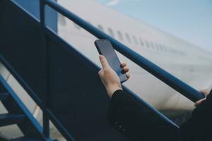 jung asiatisch Frau im International Flughafen, mit Handy, Mobiltelefon Smartphone und Überprüfung Flug beim das Flug Information Tafel foto