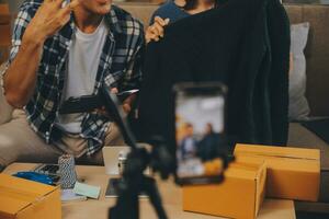 zwei asiatisch Blogger zeigen Kleider im Vorderseite von das Kamera zu Aufzeichnung vlog Video Leben Streaming beim ihr Geschäft. online Einkaufen Wagen Vorstellung. auf Sofa beim Zuhause foto