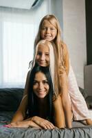 Porträt von ein glücklich Familie beim Zuhause auf das Bett. ein jung Mutter und zwei Töchter im Zuhause Kleider sind ruhen foto