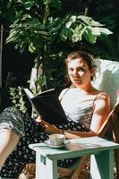 Frau, die ein Buch hält, das ernsthaft in die Kamera schaut, während sie an einem sonnigen Tag auf einem Stuhl liegt, Raum kopieren, entspannen und Hobbykonzepte foto