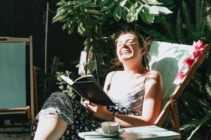 Frau lacht beim Lesen eines Buches, während sie an einem sonnigen Tag auf einem Stuhl liegt, Platz kopieren, Entspannungs- und Hobbykonzepte, soziales Netzwerk