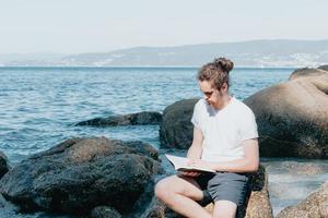 junger Mann mit langen Haaren, der an einem hellen Tag ein Buch am Strand liest, Raum kopieren, entspannen und Meditationskonzept