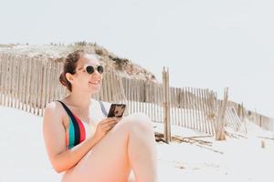 junge Frau lächelt beim Telefonieren in einem bunten Badeanzug am Strand, der ein Sonnenbad nimmt, junges Urlaubskonzept für Reisen, Kopierraum, soziales Netzwerk, Sonnenbrillen-Benutzerkonzept foto