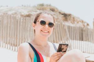 junge Frau lächelt in die Kamera, während sie am Telefon chattet und einen bunten Badeanzug am Strand trägt, der ein Sonnenbad nimmt, junges Urlaubskonzept reisen, Platz kopieren, soziales Netzwerk, Sonnenbrillen-Benutzerkonzept foto