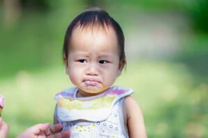 einer Jahr alt asiatisch Baby Junge macht ein ernst Ausdruck nach Essen Süss und sauer Erdbeere Eis Creme, Kind trägt ein Schürze, Kleinkind isst Süßigkeiten. foto