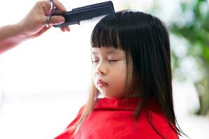 asiatisch Kind Mädchen sitzt immer noch mit ihr Augen geschlossen während Friseur ist Styling ihr Haar im bestellen zu verschönern Es. 4 Jahr alt Kind ist gehaltenen im rot Schleier zu verhindern Haar von bekommen stecken im ihr Kleidung. foto