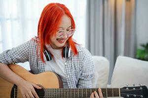 asiatisch Frau tragen Brille und Kopfhörer spielen Gitarre während Sitzung auf Sofa im das Leben Zimmer beim heim. asiatisch Frauen Schreiben Lied während spielen Gitarre beim heim. komponieren Lied Musik- Konzept. foto