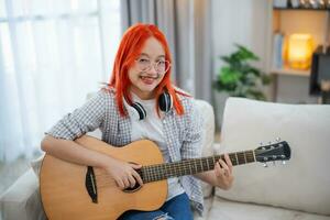 asiatisch Frau tragen Brille und Kopfhörer spielen Gitarre während Sitzung auf Sofa im das Leben Zimmer beim heim. asiatisch Frauen Schreiben Lied während spielen Gitarre beim heim. komponieren Lied Musik- Konzept. foto