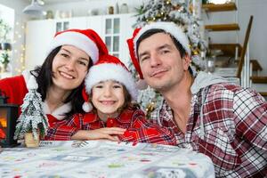 glücklich traditionell Familie von Papa, Mutter, Tochter im festlich Santa Hüte im ein Weiß Küche mit ein Weihnachten Baum und Dekor. Neu Jahr, Familie Werte. foto