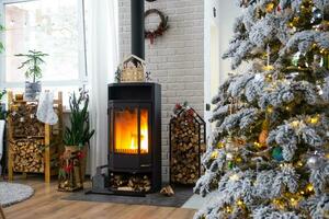 metall schwarz Herd, Verbrennung Feuerstelle Kamin im Weiß festlich Innere von Haus ist dekoriert zum Weihnachten und Neu Jahr, Weihnachten Baum. Brennholz im das Holzstapel, gemütlich und Heizung von Zuhause foto