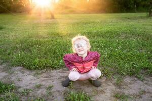 ein klein Kind runzelt die Stirn Sitzung Sitzung, hocken beim Sonnenuntergang. das Mädchen geschlossen ihr Augen mit ein Augenbinde foto