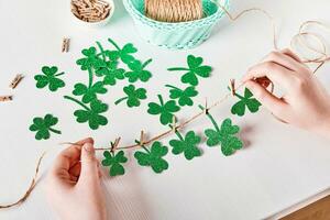 DIY St Patrick's Tag Dekor. Hände machen Kleeblätter Girlande von funkeln Papier auf Weiß Tabelle foto