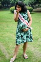 ein schwanger indisch Dame posiert zum draussen Schwangerschaft schießen und Hände auf Bauch, indisch schwanger Frau setzt ihr Hand auf ihr Bauch mit ein Mutterschaft Kleid beim Gesellschaft Park, schwanger draußen Mutterschaft schießen foto