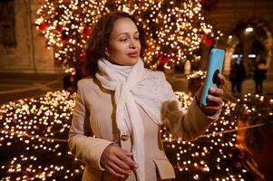 schön Frau beim Kirmes, halten Handy, Mobiltelefon Telefon, nehmen ein selbst Porträt gegen beleuchtet Weihnachten Baum im das Nacht foto