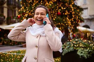 lächelnd multi ethnisch schön Frau reden auf Clever Handy, Mobiltelefon Telefon gegen beleuchtet Weihnachten Baum beim Weihnachten Kirmes foto