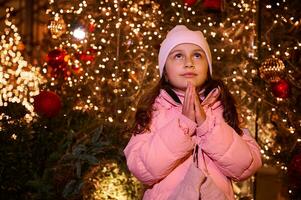 Kind Mädchen mit Hände im Gebet Geste, sieht aus hoch, macht Wunsch Stehen gegen Weihnachten Baum beim traditionell Familie Markt foto