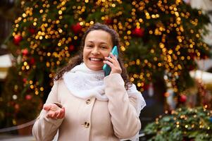 charmant multi ethnisch Frau lächelnd reden auf Clever Handy, Mobiltelefon Telefon, Stehen gegen dekoriert Weihnachten Baum beim Kirmes foto