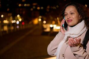 charmant jung Frau lächelnd reden auf Handy, Mobiltelefon Telefon, Stehen auf das Straße beim Nacht, mit schön Weihnachten Beleuchtung foto