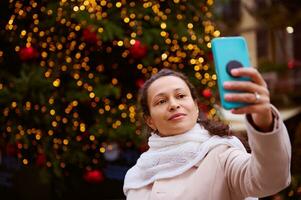 schön multi ethnisch Frau Herstellung Selfie auf ihr Smartphone, Stehen gegen beleuchtet Weihnachten Baum während Kirmes foto