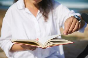 selektiv Fokus auf das Buch im das Hände von ein jung Frau Blättern durch das Seiten während lesen Roman auf das Strand foto