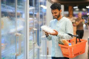 Porträt von glücklich indisch Mann Stehen im Vorderseite von das Produkt Zähler im ein Lebensmittelgeschäft speichern. Mann Kauf Lebensmittelgeschäft zum Zuhause im Supermarkt. foto