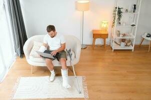 glücklich jung Mann mit Arm im ein Besetzung Sitzung auf das Couch beim Zuhause und kommunizieren auf ein Laptop foto