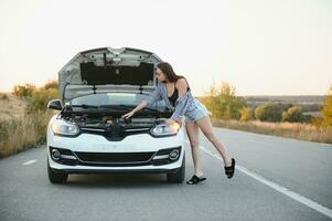 schön schlank Mädchen im Hemd und kurze Hose sieht aus im öffnen Auto Kapuze auf ein Straße foto