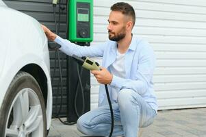 Mann halten Leistung Laden Kabel zum elektrisch Auto im draussen Auto Park. und er s gehen zu verbinden das Auto zu das Laden Bahnhof im das Parkplatz Menge in der Nähe von das Einkaufen Center. foto