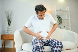 unzufrieden indisch oder arabisch Mann, sitzt auf komfortabel Sofa im gemütlich Leben Zimmer, hält seine Hände auf seine Magen, Grimassen von Schmerzen im seine Magen, leidet von Vergiftung, Krampf, Bauch Probleme. foto