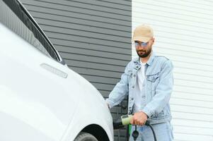 Mann halten Leistung Laden Kabel zum elektrisch Auto im draussen Auto Park. und er s gehen zu verbinden das Auto zu das Laden Bahnhof im das Parkplatz Menge in der Nähe von das Einkaufen Center. foto