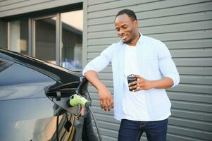 stilvoll afrikanisch Mann mit Kaffee Tasse im Hand Einsätze Stecker in das elektrisch Auto Laden Steckdose foto