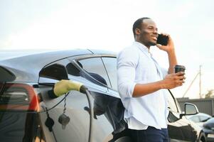 stilvoll afrikanisch Mann mit Kaffee Tasse im Hand Einsätze Stecker in das elektrisch Auto Laden Steckdose foto