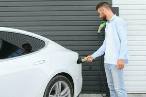 Mann halten Leistung Laden Kabel zum elektrisch Auto im draussen Auto Park. und er s gehen zu verbinden das Auto zu das Laden Bahnhof im das Parkplatz Menge in der Nähe von das Einkaufen Center. foto