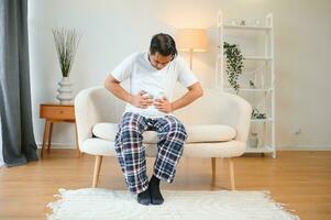 unzufrieden indisch oder arabisch Mann, sitzt auf komfortabel Sofa im gemütlich Leben Zimmer, hält seine Hände auf seine Magen, Grimassen von Schmerzen im seine Magen, leidet von Vergiftung, Krampf, Bauch Probleme. foto