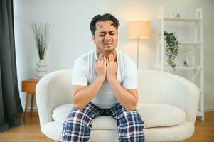 krank jung indisch Mann Leiden Kehle schmerzen während Sitzung auf Bett beim Zuhause foto