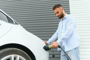 schön jung stilvoll Mann ist mit elektrisch Auto beim tagsüber Laden das Fahrzeug foto